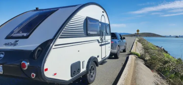 NuCamp teardrop RV rental towed by a truck on a sunny road along a bay, distant land, clear sky.. Nucamp Tab 400 2024