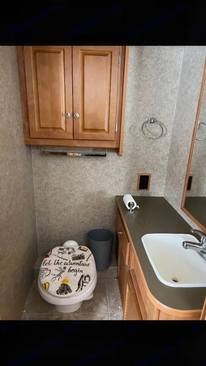 Compact RV rental bathroom featuring a sink, vanity, overhead cabinet, and adventure-themed toilet seat.. Winnebago Chalet 2010