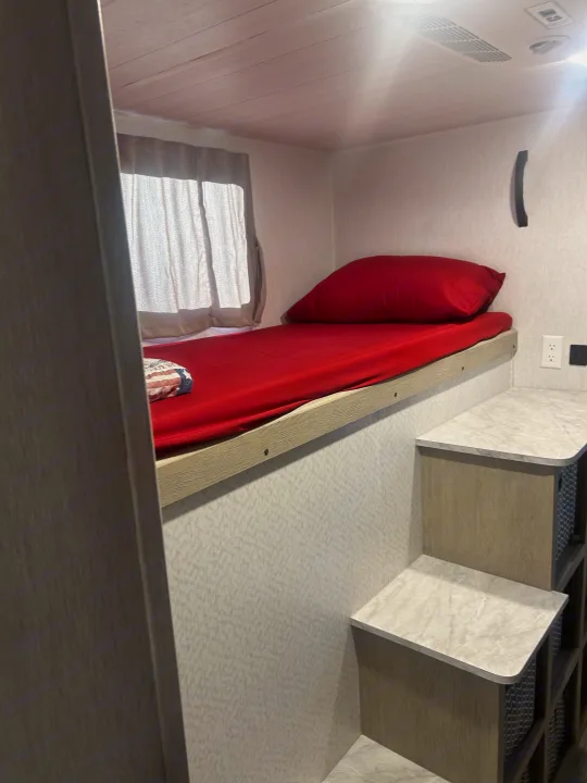 Cozy RV rental bunk bed with vibrant red bedding, a window with curtains, convenient integrated steps, and outlets for comfort.. Forest River 32BHDSX 2023