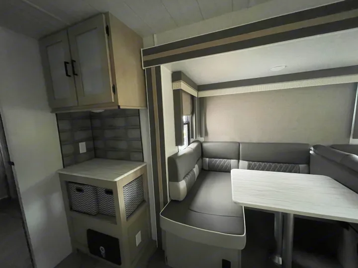 Inside a modern RV rental: a gray booth dinette with a white table, alongside a small kitchen counter with cabinets and storage.. Forest River 32BHDSX 2023
