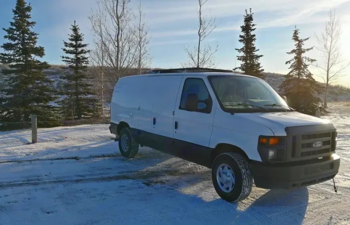 White Ford E-Series van. This RV rental is parked on a snowy road with trees and a clear sky, ready for winter trips.. Ford Ford E350 2011