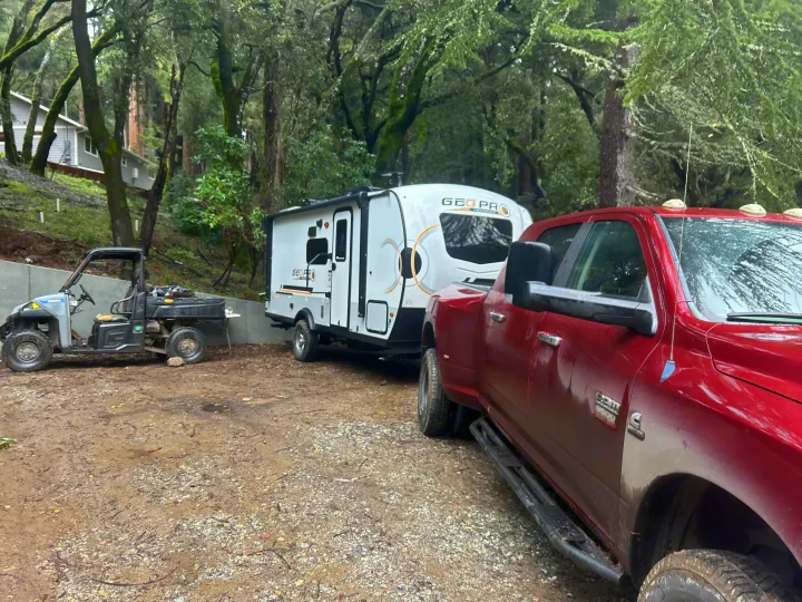 Red Ram truck towing a white Geo Pro RV rental, parked with a UTV on a dirt lot in a lush forest setting.. Geo pro Geo pro by rockwood 2022
