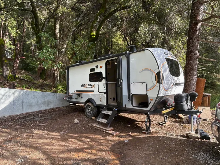 A GEO PRO RV rental is set up in a dirt camping area with a concrete wall, surrounded by lush trees.. Geo pro Geo pro by rockwood 2022