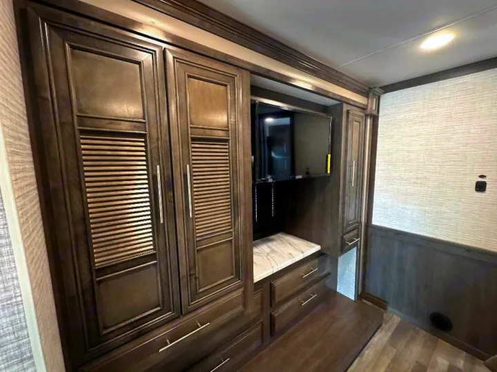 RV rental interior: dark wood cabinetry with louvered doors, a mounted TV, and dresser drawers for storage.. Entegra Coach Vision XL 2025