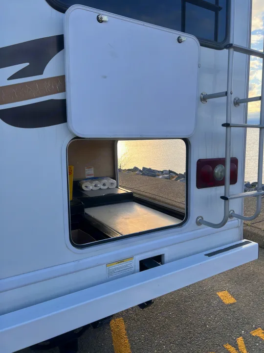 Rear of an rv rental with an open storage compartment revealing a pull-out surface and supplies, overlooking a sunny lake from a paved area.. Thor Freedom Elite 2014