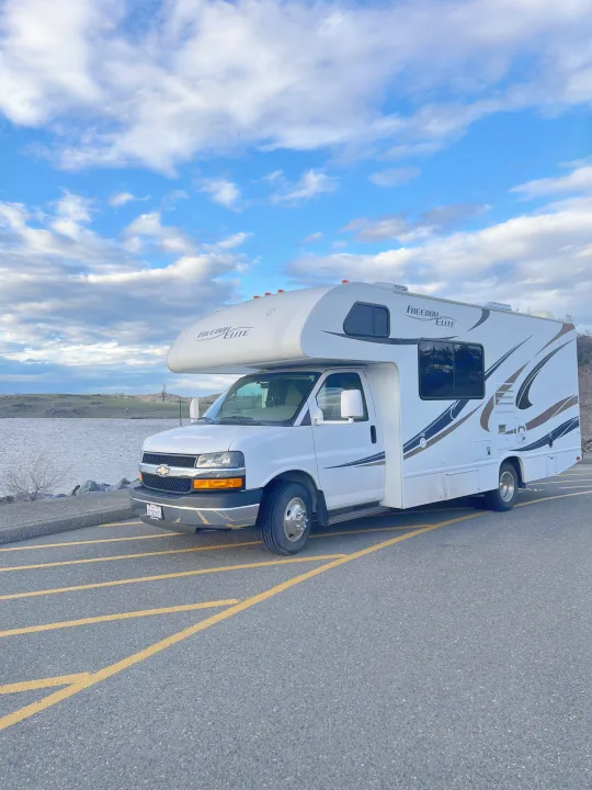 Experience the open road! This white "Freedom Elite" RV rental is parked riverside under a beautiful cloud-filled sky.. Thor Freedom Elite 2014