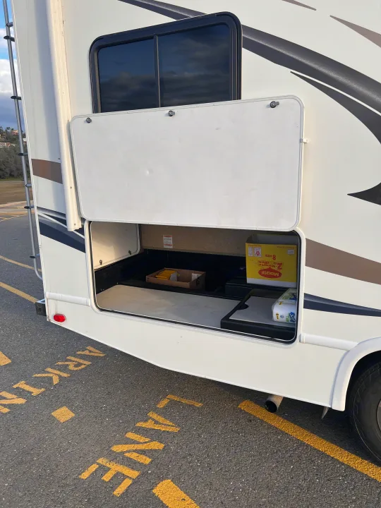 An RV rental's side storage bay is open, showing Maggi noodles, toilet paper, and essentials for the road. Parked on asphalt.. Thor Freedom Elite 2014