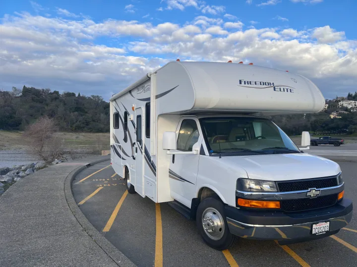 White Class C motorhome, an ideal RV rental, parked on asphalt near a lake, under a blue sky with fluffy clouds.. Thor Freedom Elite 2014
