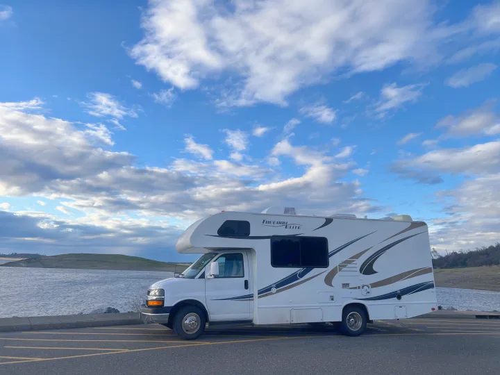 White RV rental parked by a serene lake with rolling hills under a cloudy sky, ideal for a scenic outdoor getaway.. Thor Freedom Elite 2014