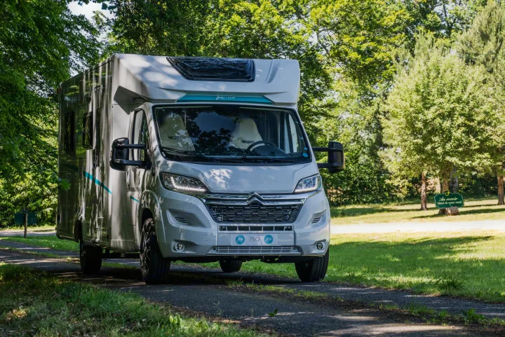 A modern white JoaCAMP 75Q RV is parked on a sunlit path, surrounded by vibrant green trees and grass. Explore rv rental options.. JOA 75Q 2024