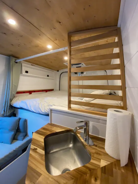Bright RV rental interior features a cozy bed, compact kitchen with sink, wooden ceiling, and slatted partition.. Nissan Other 2012