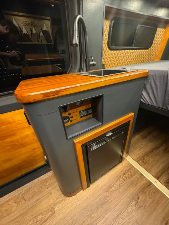 Explore this RV rental's cozy interior: a sleek kitchen area with sink, fridge, and wooden counter, next to a comfy bed.. Mercedes-Benz Sprinter 2020