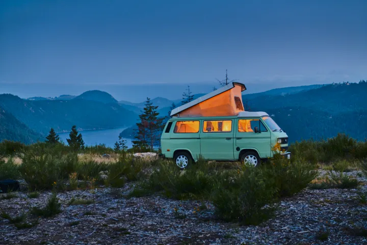 Cozy light green camper van (RV rental) parked on a mountain top at dusk overlooking a lake, illuminated inside.. Volkswagen T3 Westfalia 1986
