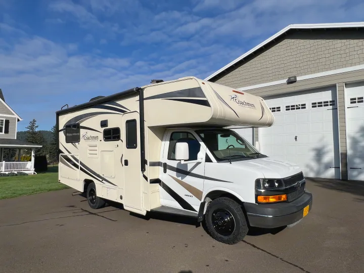 A cream Coachmen Freelander RV rental is parked in a driveway next to a house and garage, with trees and hills under a blue sky.. Forest River Coachmen Freelander 2017