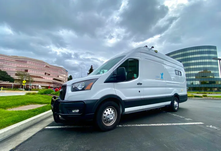 A white customized van, resembling an RV rental, is parked in a lot under a cloudy sky, with modern buildings behind it.. Ford Transit Transit 2022
