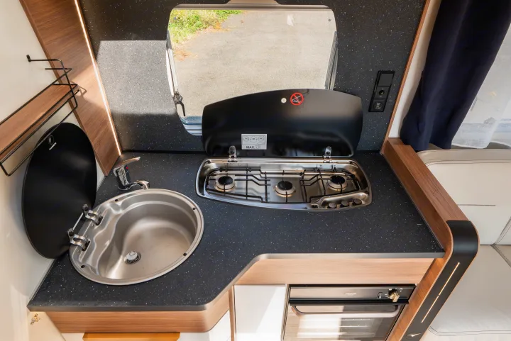 Bright rv rental kitchen with a stainless steel sink, two-burner gas stove, compact oven, and window view.. Pilote P696D ESSENTIEL 2024