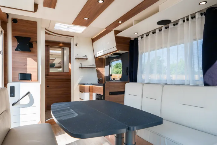 Bright RV rental interior showing a dining area with a dark table, white seating, and a compact kitchen.. Pilote P696D ESSENTIEL 2024