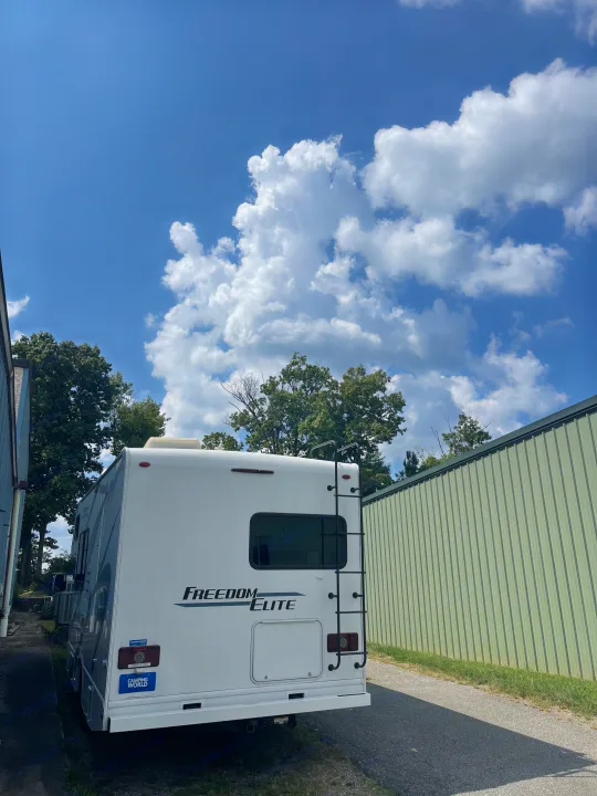 A white Freedom Elite RV, possibly an RV rental, is parked between a blue building and a green metal wall on a sunny day.. Thor Freedom Elite 2021