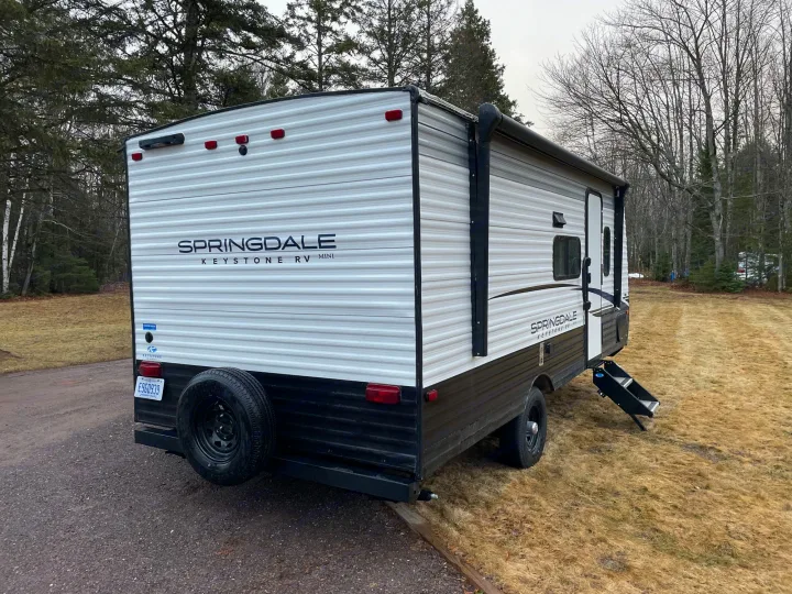 A white and black Springdale Keystone RV Mini travel trailer, ideal for an RV rental, parked on grass with steps down.. Keystone RV Springdale 2023