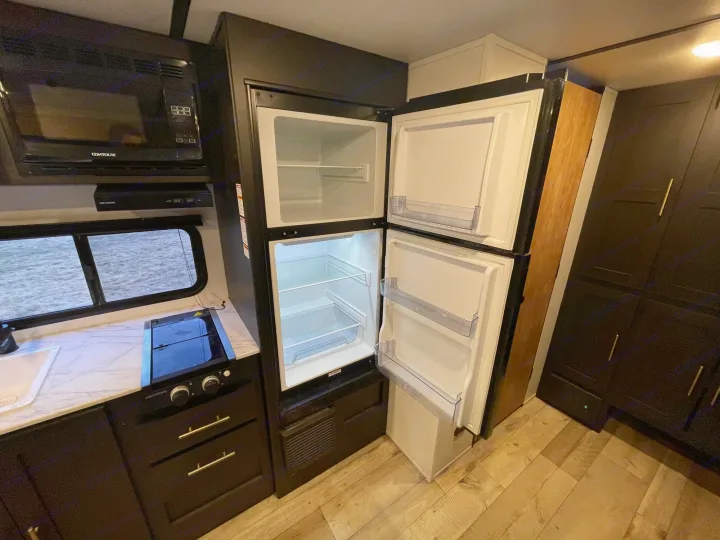 Inside an RV rental: a compact kitchen featuring a microwave, open two-door fridge, stove, sink, and dark cabinetry.. Keystone RV Springdale 2023