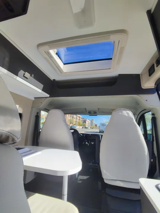 Bright RV rental interior with large skylight, light seats, and dining table facing the road.. McLouis Menfys Van 3 Maxi 2024