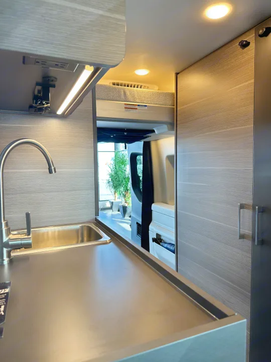 Modern rv rental interior: compact kitchen with a stainless steel sink, dark counter, LED lights, and a rear sleeping area.. Mercedes-Benz Entegra Launch 2024
