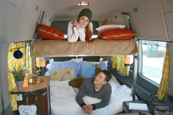 Cozy RV rental interior shows a woman on an upper bunk and a man on the lower bed inside a custom camper van.. Ford Transit 2023