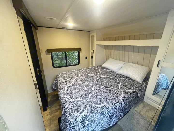 Cozy rv rental bedroom with a patterned bed, two pillows, built-in storage, and a window offering a view of trees.. Keystone RV Hideout 2022
