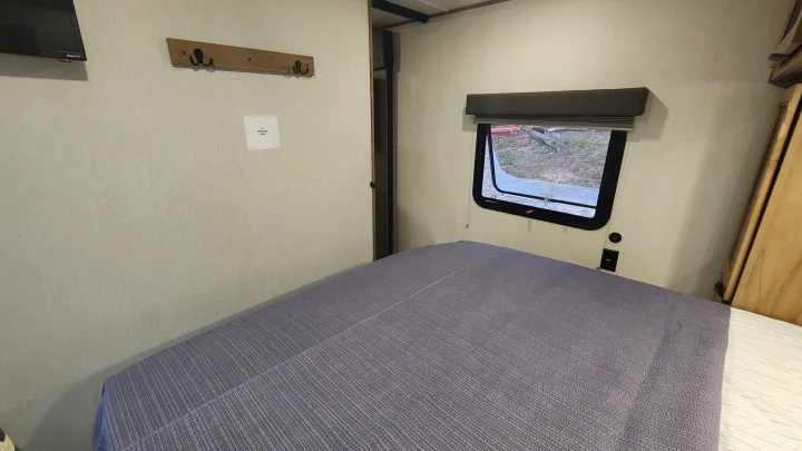 Inside an RV rental: a cozy bedroom with a neatly made bed, window view, TV, and wall-mounted coat hooks.. Forest River 22sw-l 2024