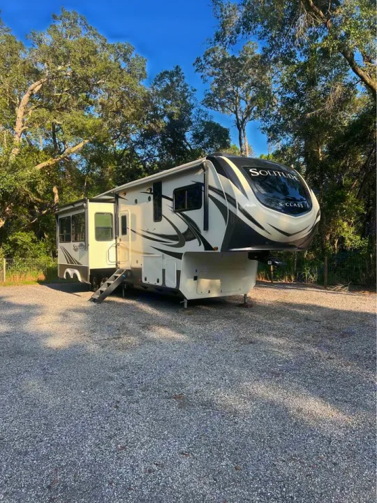 Experience nature! This beautiful Solitude fifth-wheel RV rental is parked on a gravel lot, surrounded by lush trees & a blue sky.. Grand Design Solitude 2020