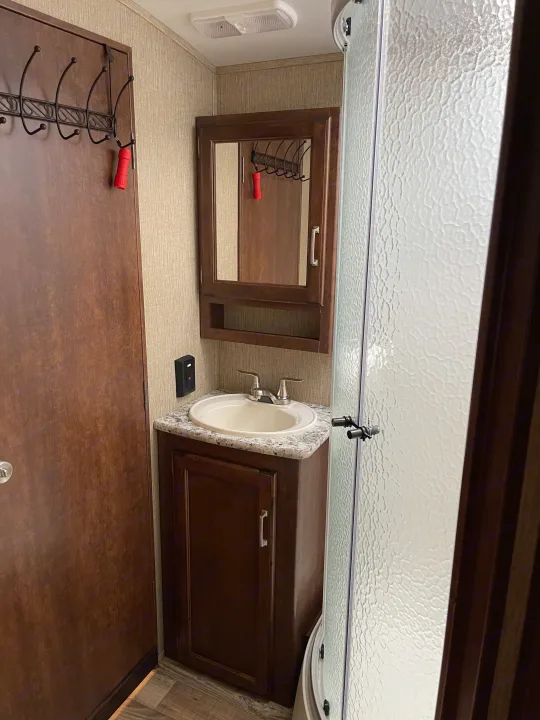 RV rental bathroom interior showing sink, vanity, mirrored cabinet, shower, and a door with a coat rack & red flashlight.. Keystone RV Outback Super-Lite 2018