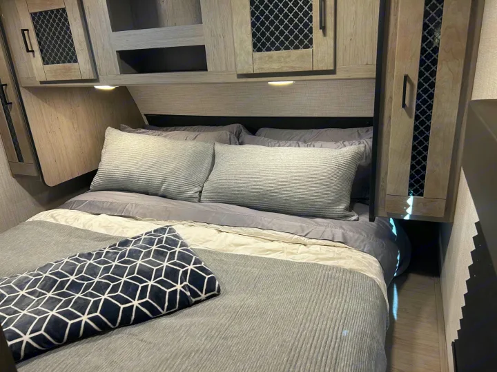 Cozy gray bedroom in an RV rental, featuring overhead cabinets, soft bedding, and integrated lighting. Ideal for travel.. Heartland RVs Mallard 2021