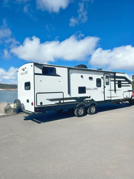 White Mallard travel trailer, ideal for an RV rental, parked by a scenic lake under blue skies.. Heartland RVs Mallard 2021