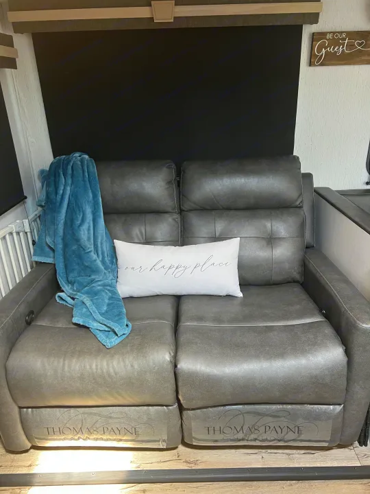 This gray two-seat recliner sofa with a blue blanket and "our happy place" pillow provides comfortable seating for an RV rental.. Keystone RV Astoria 2903BH 2022