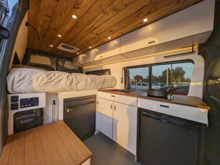 Step inside this modern RV rental featuring a cozy bed, a functional kitchen with sink & cooktop, white cabinets, and a warm wood ceiling with recessed lighting.. Mercedes-Benz Sprinter 2023