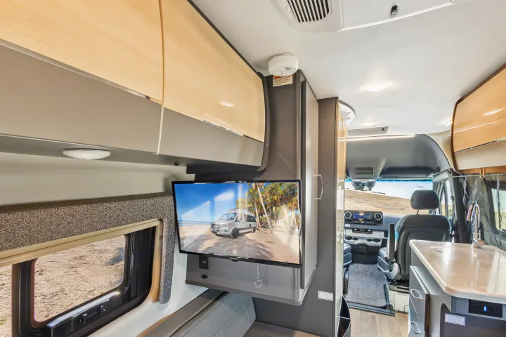 RV rental interior: a cozy living area with a TV displaying a beach scene, overhead storage, and the driver's cabin.. Thor Sanctuary 2023