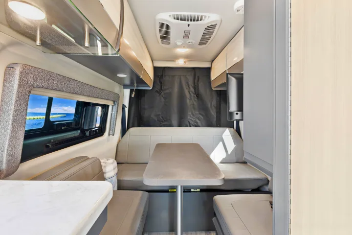 Modern RV rental interior: dinette seating, overhead storage, AC, and a scenic ocean view from the window.. Thor Sanctuary 2023