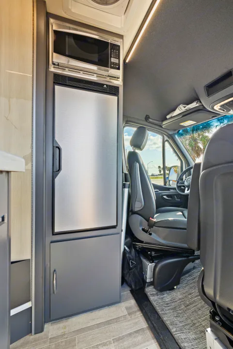 Inside this rv rental camper van: stainless steel microwave, large refrigerator, and front seats with a scenic road view.. Thor Sanctuary 2023