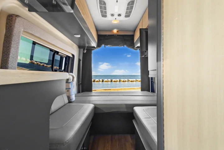 Step inside this RV rental! View of a modern camper van interior, featuring grey seating, wood-grain accents, and a large ocean view.. Thor Sanctuary 2023