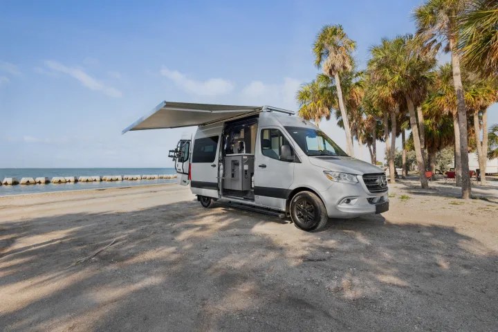 This silver Mercedes Sprinter camper van RV rental is parked on a sandy beach near palm trees and the ocean.. Thor Sanctuary 2023