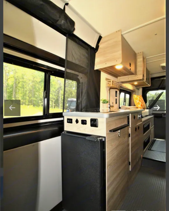 This RV rental interior features a compact kitchen with a sink, refrigerator, wood cabinets, and large windows overlooking trees.. Winnebago Solis 2023