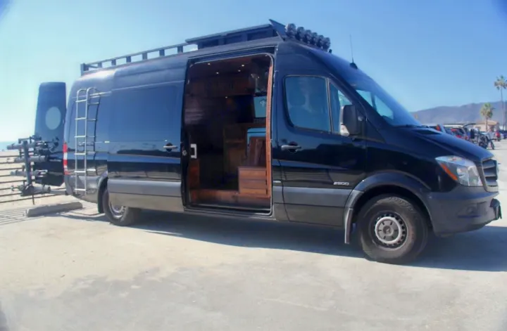 A sleek black custom Sprinter van RV rental with open door revealing warm wooden interior. Equipped for road trip adventures by the beach.. Mercedes-Benz Sprinter 2016