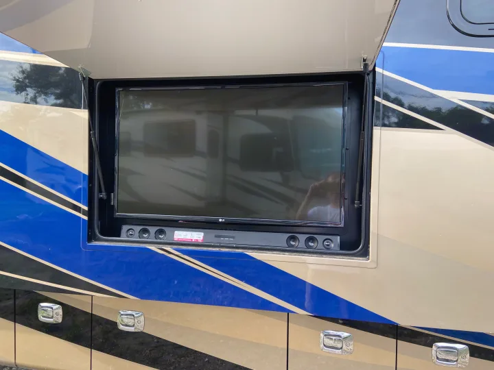 RV rental's side with blue, black, and beige stripes. An open compartment reveals an outdoor TV and soundbar.. Tiffin Motorhomes Allegro Bus 2017
