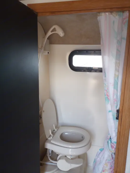 Compact RV rental bathroom: white toilet, wall-mounted shower head, small window, and floral shower curtain.. Scamp Scamp Trailer 2011