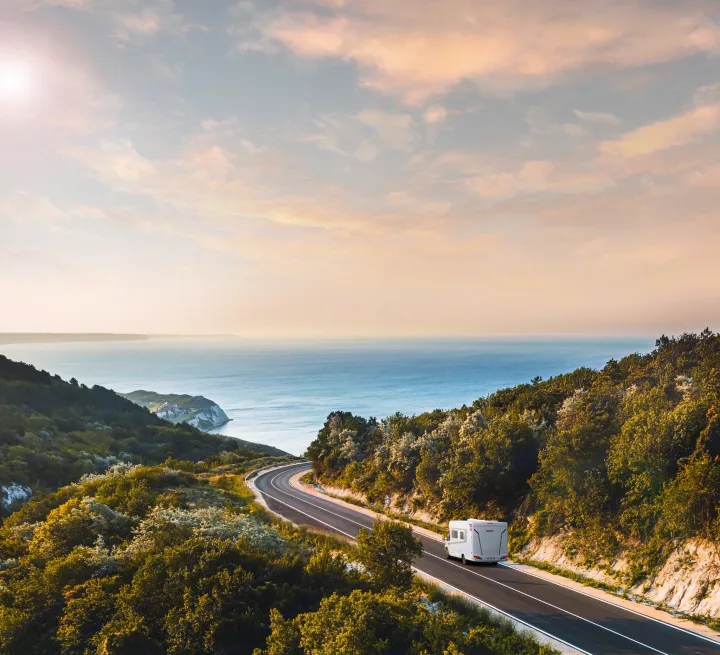 A white RV drives along a scenic coastal road overlooking the ocean and lush green hills, perfect for an RV rental adventure.. Pilote P696D ESSENTIEL 2024