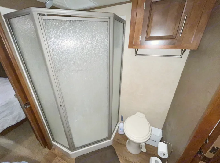 This RV rental bathroom features a compact corner shower with textured glass, a toilet, and an overhead wooden cabinet, all on a wood-look floor.. Forest River XLR Thunderbolt Toy Hauler 2015