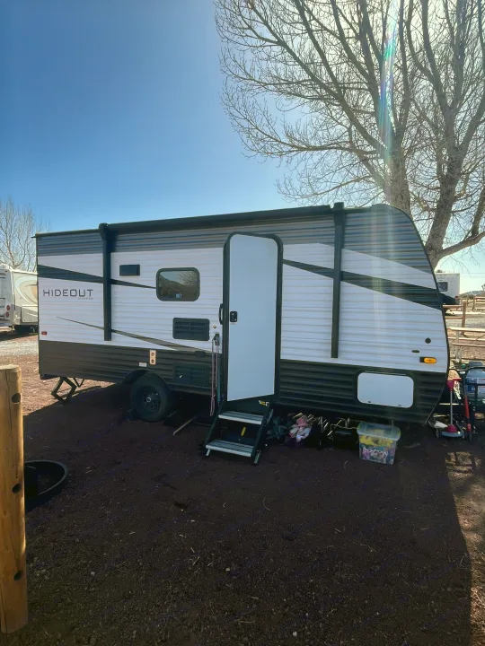 A white and black Hideout travel trailer (RV rental) is parked on a dirt lot at a sunny campsite with steps down and door open.. Keystone RV Hideout 2021
