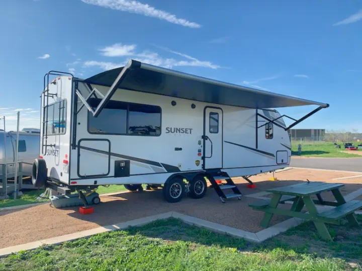 A white Sunset travel trailer RV rental parked at a sunny campsite with its awning extended, alongside a picnic table.. Crossroads RV Sunset Trail Super Lite 2022