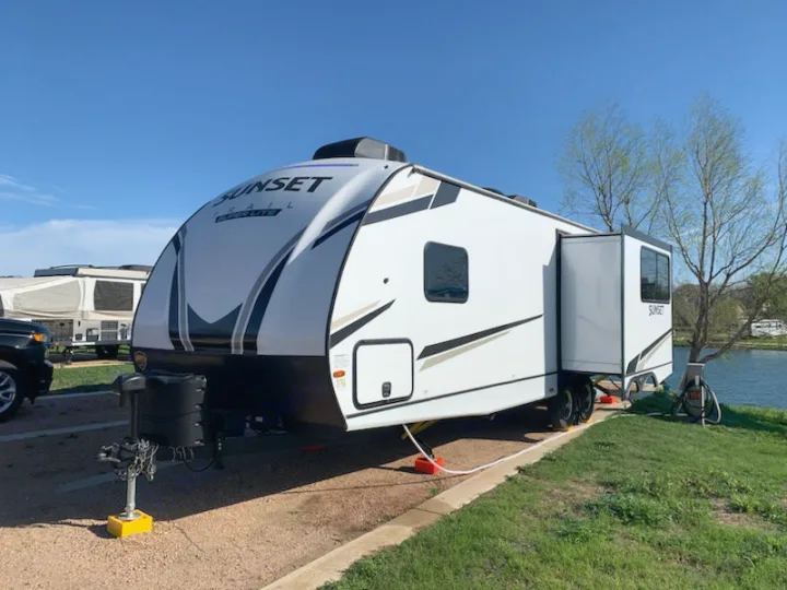 An RV rental: a white travel trailer, fully set up on a gravel campsite by a lake, ready for adventure.. Crossroads RV Sunset Trail Super Lite 2022