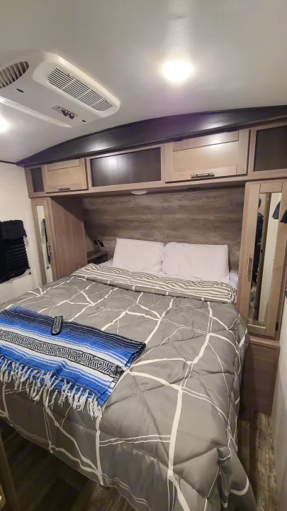 View inside an RV rental's cozy bedroom, showcasing a bed with gray comforter, white pillows, and overhead storage/AC unit.. Crossroads RV Sunset Trail Super Lite 2022